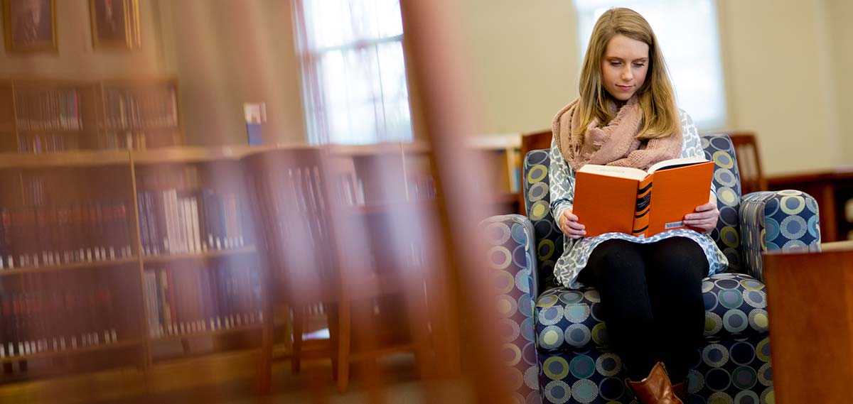 Female student sits on an armchair reading a book in the Leland Speed Library on Mississippi College's campus.
